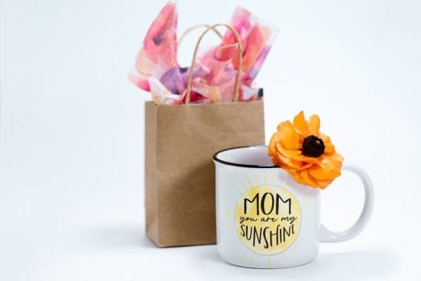 A mug with 'Mom you are my sunshine' beside a gift bag with colorful tissue paper, perfect for Mother's Day.